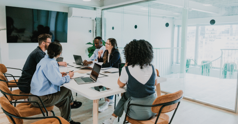 A diverse team of people sitting around a table in a bright, modern office, chatting and collaborating. This represents building a team for organisational change.