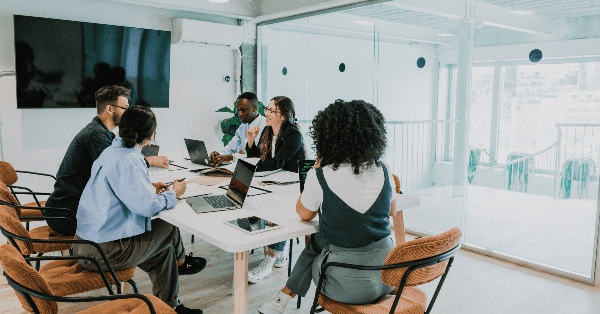 A diverse team of people sitting around a table in a bright, modern office, chatting and collaborating. This represents building a team for organisational change.