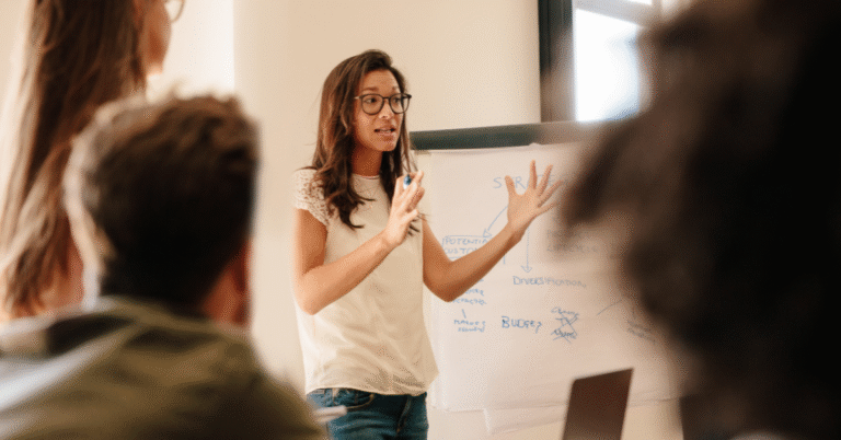 A woman wearing glasses and a white shirt stands in front of a whiteboard, gesturing with her hands while speaking to an audience in a presentation setting. This visual represents a Great Leader in 2025 actively engaging and communicating.