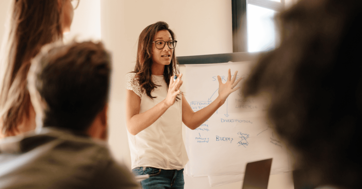 A woman wearing glasses and a white shirt stands in front of a whiteboard, gesturing with her hands while speaking to an audience in a presentation setting. This visual represents a Great Leader in 2025 actively engaging and communicating.