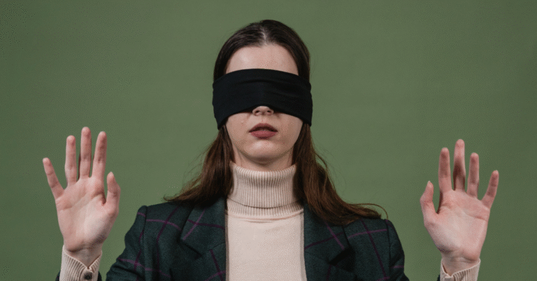 A person with long, dark hair wearing a black blindfold, a light-colored turtleneck, and a patterned blazer stands against a green background, holding both hands up with palms facing forward as if feeling their way or signalling a halt. This image visually represents leadership blind spots.