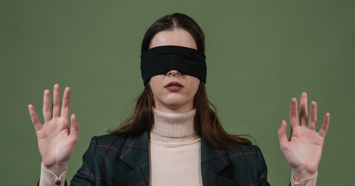 A person with long, dark hair wearing a black blindfold, a light-colored turtleneck, and a patterned blazer stands against a green background, holding both hands up with palms facing forward as if feeling their way or signalling a halt. This image visually represents leadership blind spots.