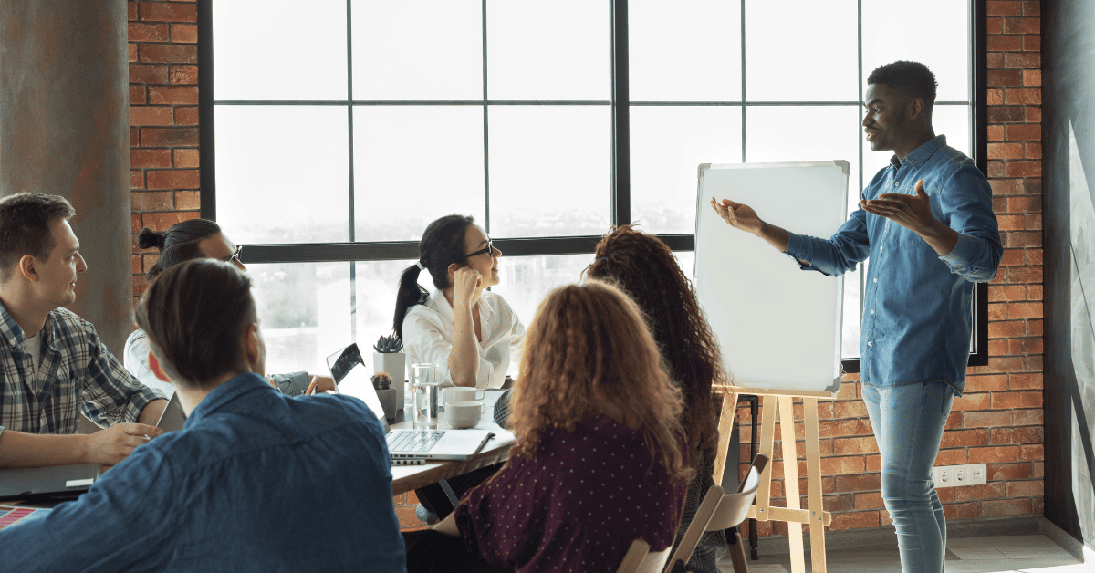 A male presenter in a denim shirt stands beside a whiteboard, gesturing to a diverse group of attentive colleagues seated around a conference table in a modern, brick-walled office with large windows. This image represents the active guidance seen in coaching vs managing.